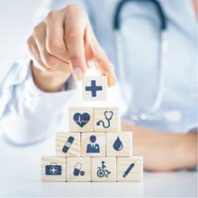 decorative image of a medical professional wearing a white coat and stethoscope stacks wooden blocks printed with healthcare icons, such as a cross, heart monitor, stethoscope, pill, doctor, blood drop, medical kit, wheelchair, and syringe, into a pyramid